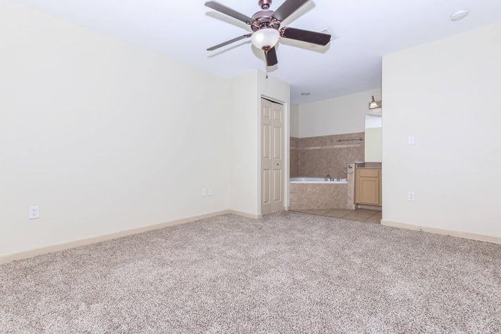 Empty room with light beige walls, a ceiling fan, and carpeted floor. A doorway leads to a bathroom area featuring a bathtub and a small vanity. Natural light comes from an unseen window, giving a spacious and clean appearance.