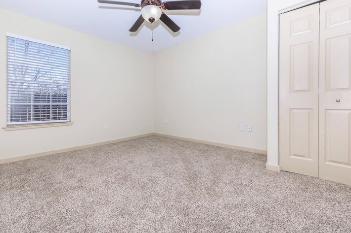 A spacious, empty room with light beige walls and carpet. It features a ceiling fan, a window with white blinds allowing natural light to enter, and a closet with double doors. The room is tidy and ready for furnishing, creating a warm and inviting atmosphere.