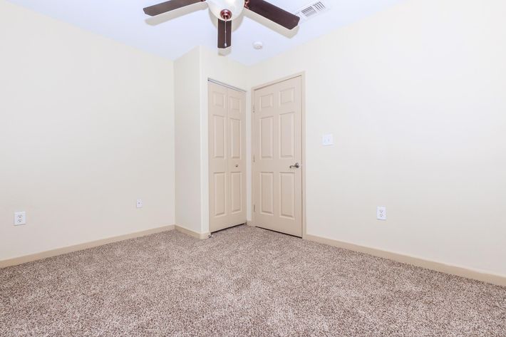 A sparsely furnished room featuring beige walls and a light carpeted floor. There are two closed beige doors on the left and a ceiling fan with wooden blades overhead. The room has a neutral appearance, ideal for various uses, such as a bedroom or office.