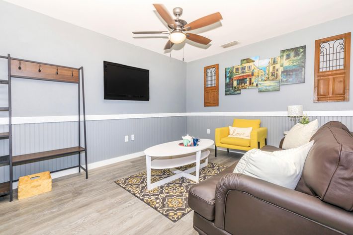 A cozy living room featuring a brown leather sofa with decorative pillows, a round white coffee table, a television mounted on the wall, and a stylish rug. The walls are painted gray with white wainscoting. There is a yellow chair with the word "Love," a small shelf for storage, and artwork depicting a quaint scene.