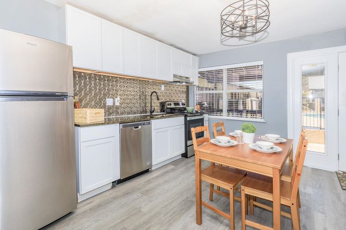 Modern kitchen featuring stainless steel appliances, including a refrigerator and dishwasher. The cabinetry is white with a stylish backsplash. A wooden dining table with four chairs is set for a meal, and a small potted plant adds a decorative touch. Natural light streams in through a window.