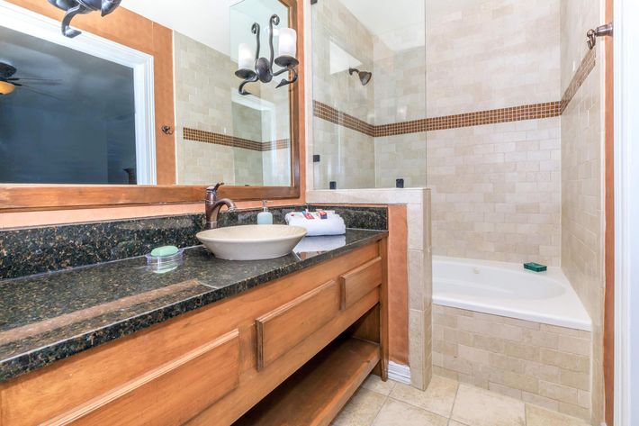 A spacious bathroom featuring a wooden vanity with a dark granite countertop, a modern sink, and a large mirror above. To the side is a relaxing bathtub, complemented by tiled walls and a neutral color palette. Soft lighting enhances the inviting atmosphere.