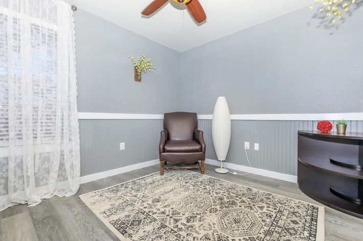 A cozy, minimally furnished room featuring a brown leather armchair, a tall white floor lamp, and a dark wood cabinet. The walls are painted in a soft gray, with white wainscoting, and a textured area rug lies on the floor. Natural light filters through a sheer white curtain on the window, enhancing the inviting atmosphere.