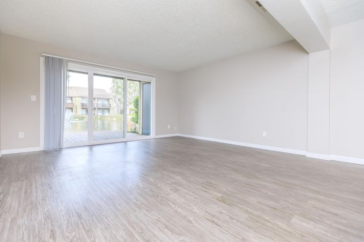 Spacious empty living room with light wood laminate flooring, large sliding glass doors leading to a balcony overlooking a serene water view. The walls are painted in a neutral color, and there are no furniture or decorations, emphasizing the openness of the space. Natural light floods the area, creating a bright ambiance.