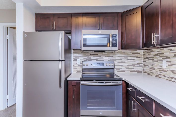 Modern kitchen featuring dark wood cabinetry, stainless steel refrigerator and oven, and a microwave above the stove. The backsplash is made of small, rectangular tiles in neutral tones, complementing the sleek countertops. The space is well-lit and organized, creating a functional cooking area.