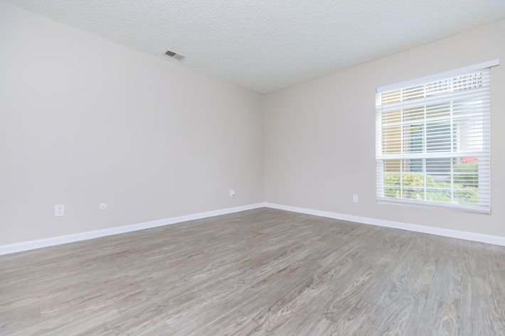 Empty room with light beige walls and wooden flooring. A single window with white blinds allows natural light to enter, illuminating the space. The room is spacious with no furniture, creating a minimalistic and clean appearance.