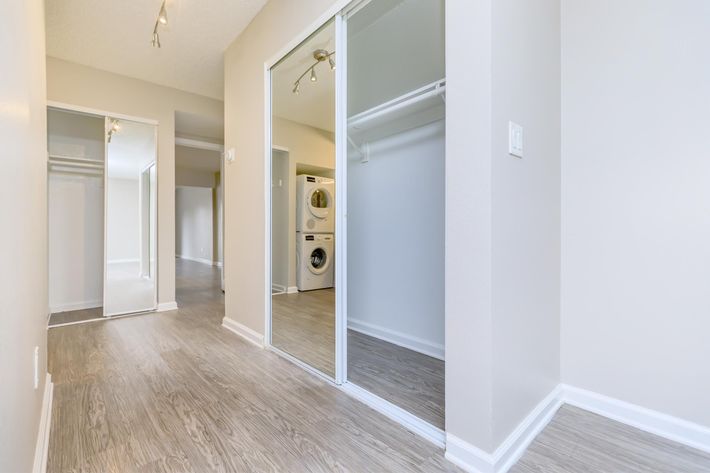 A well-lit interior hallway showing a laundry area with a washing machine and dryer. On the left, there's a closet with sliding mirrors, and the floor is covered in light-colored laminate. The walls are painted in a neutral tone, contributing to a spacious and airy feel.