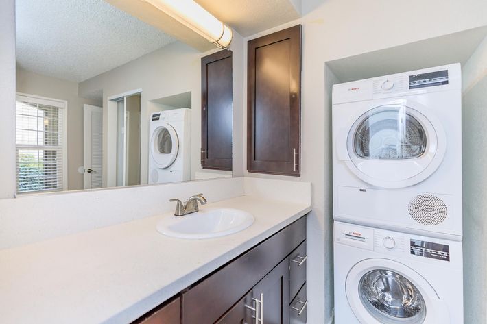 A modern laundry area with a stacked washer and dryer next to a countertop sink. The wall is painted light, and there are dark wood cabinets above the sink. In the background, a doorway leads to another room, and a window provides natural light. The space is clean and well-organized.