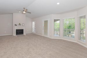 Spacious living room featuring light-colored walls, a ceiling fan, and a cozy fireplace. Large windows with white blinds allow natural light to fill the room, while plush carpet flooring adds warmth. The area is bright and inviting, with access to outdoor views.