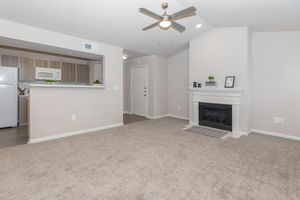 A spacious living area featuring beige walls and carpet. There is a ceiling fan, a white fireplace with a gray mantle, and an open kitchen area with wooden cabinetry. A door leads to the entrance, with a small decorative shelf on the side. Natural light fills the room, creating an inviting atmosphere.