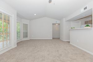 Spacious, light-filled living area with beige carpet and neutral wall color. Features large windows with blinds, a ceiling fan, and a kitchen bar area. Open layout leading to a hallway, creating a welcoming atmosphere suitable for various uses.