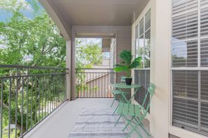 A sunny balcony with a small green table and two chairs, adorned with a potted plant. The space features a light-colored rug and has views of surrounding trees and buildings, creating a relaxing outdoor atmosphere.
