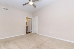 A spacious, empty room with light beige walls and tan carpet. A ceiling fan is visible, and there is an open doorway leading to a bathroom or another room. The overall setting is minimalistic and well-lit, creating an inviting atmosphere.