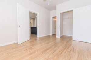 A spacious, empty room featuring light-colored walls and hardwood flooring. It has two open doors leading to separate areas, one with a closet space and another visible doorway. Natural light fills the space, creating an airy atmosphere, with a ceiling fan visible in the upper part of the room.