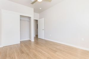 A spacious, empty room featuring light-colored walls and wooden flooring. There is a closed white door on the right, a closet with sliding doors on the left, and a ceiling fan mounted overhead. The room is well-lit, giving it a bright and airy feel.