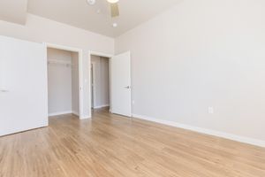 A spacious, empty room with light-colored walls and a warm wooden floor. Two doors are visible: one leading to a closet and the other to a hallway. A ceiling fan is installed, and the overall ambiance is bright and inviting, suitable for a living or bedroom space.