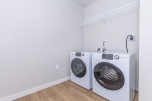 A modern laundry room featuring a pair of white front-loading washing machines side by side. The machines are placed on a wooden floor, and there is a shelf above them for storage. The walls are painted in a light color, creating a clean and bright space.