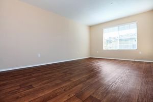 A spacious, empty room with light brown wooden flooring and a large window. The walls are painted a soft beige, and there are white baseboards. Natural light streams in through the window, illuminating the uncluttered space.