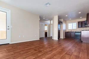 An interior view of a modern, empty living space featuring wooden floors, light-colored walls, and natural light from a window. The kitchen area includes dark cabinetry and stainless-steel appliances, while a door leads outside. There are also doorways leading to other rooms, indicating a spacious layout.