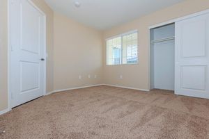 An empty room with beige walls and carpeted flooring. A window with blinds allows natural light in. There is a closet with sliding doors on the right, and a white door is visible on the left side of the room. The space is clean and unfurnished, creating a blank canvas for potential decoration.