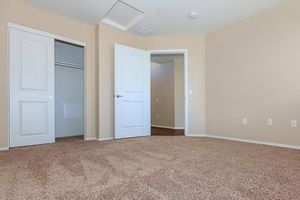 A spacious, empty room with light beige walls and carpeted flooring. Two doors are visible: one leading to a closet and the other to an adjacent room. Natural light brightens the space. The overall atmosphere is clean and uncluttered, ideal for potential furnishing and personalization.