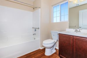 A clean, well-lit bathroom featuring a white bathtub with a shower, a modern toilet, and a double sink vanity with dark wood cabinetry. The room has light beige walls and a window allowing natural light to enter, enhancing the bright atmosphere.
