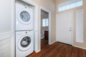 A clean and bright laundry area featuring a stacked washer and dryer. To the right, a doorway leads to a bathroom with a toilet and a window, while the front door is visible on the left. The walls are painted a soft beige, and there are wooden floors with natural light streaming in from the windows.