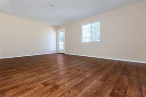 An interior view of a spacious room with light-colored walls and a wooden floor. There is a window allowing natural light onto the floor and a door leading outside. The room appears empty and inviting, suggesting potential for various furnishings or activities.