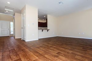 A spacious interior of an empty room featuring light-colored walls and a hardwood floor. The room includes a doorway leading to a hallway and an opening to a kitchen with dark wood cabinetry in the background. Natural light enters through a window, creating a bright atmosphere.