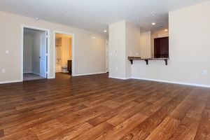 A spacious, empty room with hardwood flooring and light-colored walls. There are open doorways leading to another room and a bathroom. A small kitchen area with a bar counter is visible in the background, featuring dark cabinets. Natural light enters the space, creating a bright and inviting atmosphere.