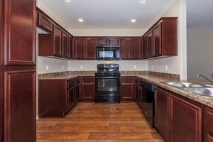 A modern kitchen featuring dark wood cabinetry, a black microwave, and an oven. The countertops are a light brown laminate, and there are dual sinks next to a window. The floor is hardwood, and the walls are painted in a light neutral color. The kitchen is well-lit with overhead lights.