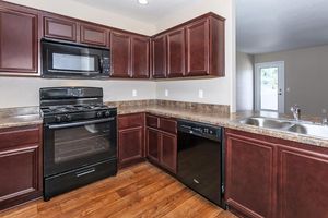 Modern kitchen featuring dark wood cabinetry, a black microwave, gas stove, and dishwasher. Countertops are a light brown stone, complemented by a wood floor. Natural light enters from a nearby window, enhancing the inviting atmosphere.