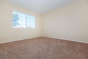 A spacious, empty room with beige walls and a light carpeted floor. Natural light streams through a window with white blinds, offering a view of greenery outside. The room features minimal decor, creating a blank canvas for personalization.