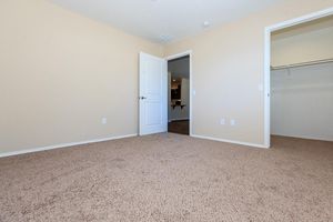 A spacious, empty bedroom with beige walls and carpet. There is a closed white door leading to another room and an open doorway leading to a closet. The room is well-lit with natural light, creating a bright and inviting atmosphere. No furniture is present, emphasizing the room's potential for personalization.