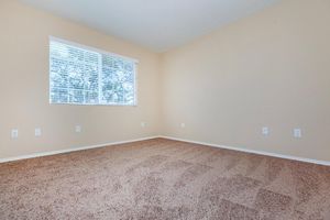 Empty room with beige walls and light brown carpet. A large window with white blinds allows natural light to enter, illuminating the space. The room features several electrical outlets and is free of furniture, creating an open and airy atmosphere.
