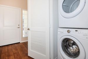 A corner of a room featuring a stacked washer and dryer unit beside a closed white door, with a window partially visible on the left. The floor is hardwood, and the walls are painted light colors, creating a bright and tidy laundry space.