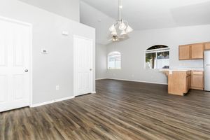 A spacious, open-concept living area with light-colored walls and wood-like flooring. There are two doors leading to adjacent rooms, a large window with blinds allowing natural light, and a ceiling fan with pendant lighting. A kitchen area with wood cabinets is partially visible in the background.