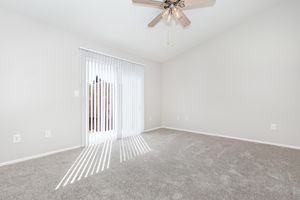 A spacious, empty room with light gray walls and carpeted flooring. It features a ceiling fan with light fixtures and a sliding glass door covered by vertical blinds, allowing natural light to stream in, casting shadows on the floor. The overall atmosphere is bright and airy.