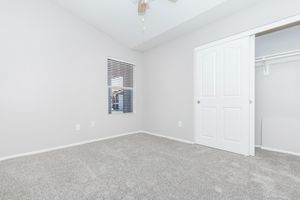 A spacious, empty bedroom featuring light gray walls and plush carpet. There is a small window with blinds on the left side, allowing natural light to enter. A closed white door on the right leads to a closet. The room has a ceiling fan and minimal furnishings, creating a clean and open atmosphere.