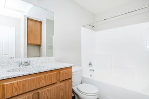 A bright and clean bathroom featuring a white bathtub and shower combination, a white toilet, and a wooden vanity with a speckled countertop. The walls are painted in a light color, and there is a large mirror above the sink area. A cabinet is visible in the background. Natural light comes from a window above.