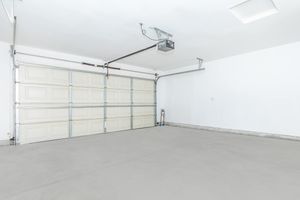 A clean and empty garage featuring a white wall, a large overhead door with a beige panel design, a ceiling light fixture, and a garage door opener mounted on the wall. The floor is smooth and concrete, with no items or furniture present.