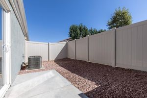 A view of a small outdoor area featuring a gray wall, a gravel surface, and an air conditioning unit. The sky is clear and blue, with a few trees visible in the background. The space is enclosed with a light-colored fence, creating a private, secluded atmosphere.