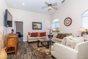A cozy living room featuring beige sofas with colorful cushions, a glass coffee table, and a decorative area rug. A large wall clock and a mirror are mounted on the walls, with windows allowing natural light in. There are lamps beside the sofas, and a wooden cabinet adds warmth to the space.