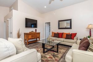 A comfortable living room featuring cream-colored sofas, a glass coffee table, and a decorative area rug. There is a wall-mounted TV, a wooden side table, and a doorway leading to another room. Natural light enters through a window, and a decorative plant adds a touch of greenery to the space.
