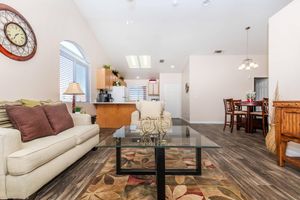 A spacious living area featuring a cream-colored sofa with decorative pillows, a glass coffee table with a floral pattern rug beneath, a wooden dining table set for four, a modern kitchen in the background, and large windows allowing natural light to fill the room.
