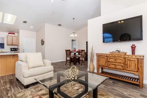 A cozy living room featuring a light-colored armchair, a glass coffee table with a decorative centerpiece, and a wooden sideboard. The space is well-lit with natural light, has a wall-mounted TV, and an adjacent dining area with a wooden table and chairs. The flooring is polished wood.