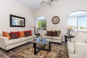 A cozy living room featuring a cream-colored sofa adorned with orange and brown pillows, a glass coffee table with decorative items, two lamps, and a large wall clock. Sunlight streams through arched windows, highlighting the warm, inviting atmosphere.