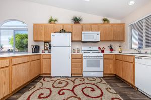 A modern kitchen with light wood cabinets, a white refrigerator, a gas stove, and a microwave. The countertop is clear with a few decorative items. A window lets in natural light, and there's a patterned red and cream rug on the floor. Green plants are placed on top of the cabinets.