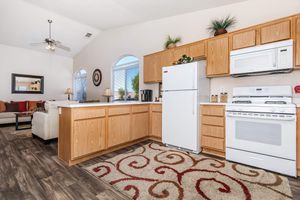 A modern kitchen featuring light wooden cabinets, a white refrigerator, and a white stove with an oven. The room has large arched windows allowing natural light, a ceiling fan, and a decorative rug with swirling patterns on the floor. There is a small dining area visible with a sofa in the background.