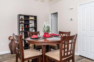 A cozy dining area featuring a round wooden table set with red napkins, a decorative centerpiece of a small artificial tree, and dark wooden chairs. In the background, a black shelving unit displays decorative items, and a doorway leads to another room. The walls are a soft beige color.